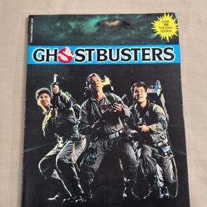 May include: A book cover featuring a photo of the Ghostbusters from the 1984 film. The cover is blue and white with the title "Ghostbusters" in large, white letters. The text "A Storybook by Anne Digby" and "Based on the smash hit film" is printed below the title.