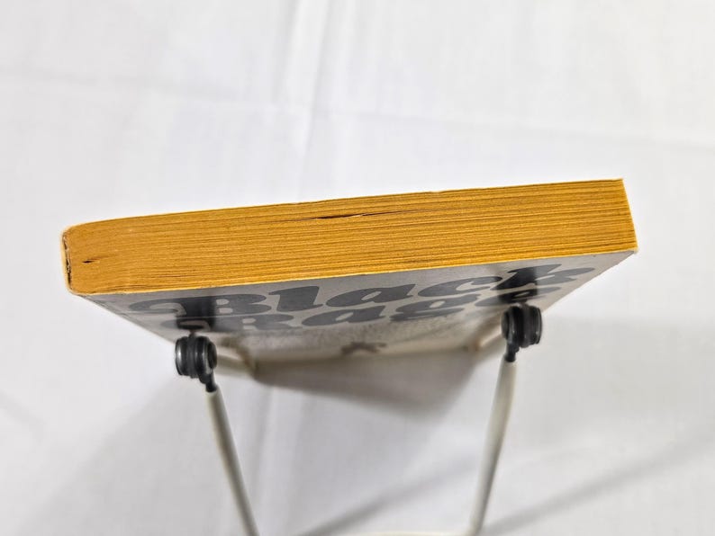 May include: A book titled "Black Rage" with yellowed pages and a black and white cover. The book is propped up on a white metal stand against a white background. The book's edges are a golden yellow color.