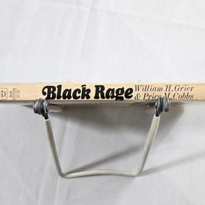 May include: A vintage book titled "Black Rage" by William H. Grier & Price M. Cobbs. The book has a light tan cover with black lettering and is held by a white metal book stand. The book is a non-fiction work.