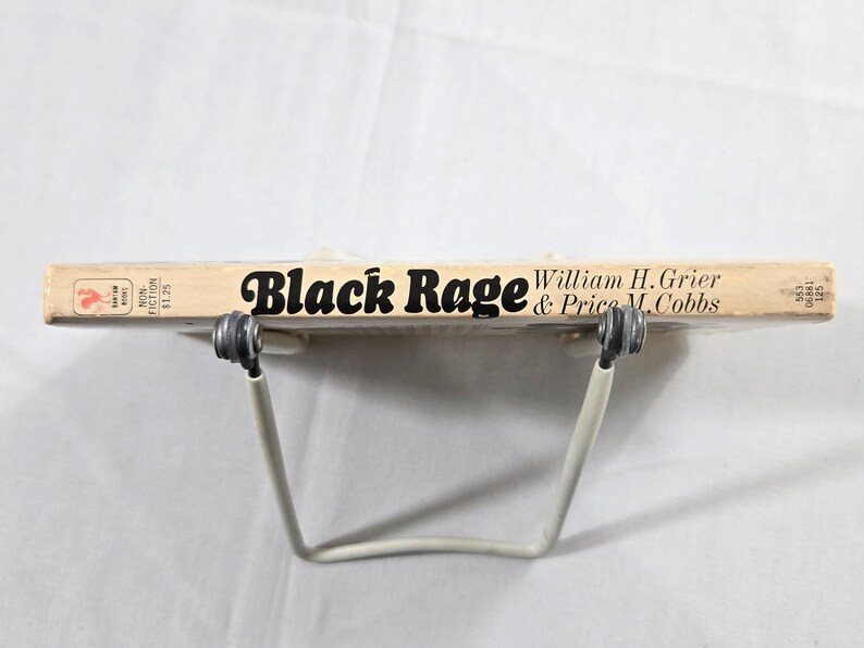 May include: A vintage book titled "Black Rage" by William H. Grier & Price M. Cobbs. The book has a light tan cover with black lettering and is held by a white metal book stand. The book is a non-fiction work.
