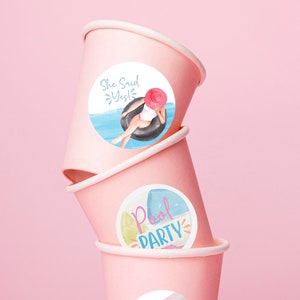 Puede incluir: Tres vasos de papel rosa con pegatinas redondas. La pegatina superior dice "She Said Yes!" y presenta a una mujer en traje de baño flotando en un flotador de piscina. La pegatina del medio dice "Pool Party" y presenta un flotador de piscina y palmeras. La pegatina inferior dice "Rachel and Ben" y presenta un anillo de diamantes y un flotador de piscina.