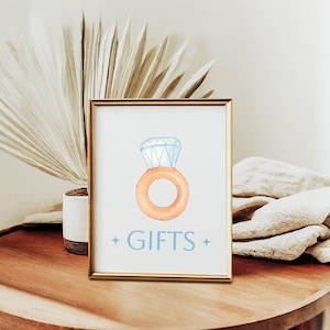 Peut inclure: Une pancarte dorée encadrée avec une illustration aquarelle d'une bague surmontée d'un gros diamant. La bague est orange et le diamant est bleu. La pancarte indique "GIFTS" en texte bleu.