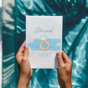 Puede incluir: Una tarjeta blanca con un fondo de acuarela azul y una ilustración de dibujos animados de un anillo con un diamante. El texto en la tarjeta dice "She said YES!"