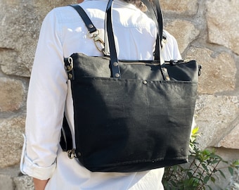 Puede incluir: Una mochila de lona negra con cierre de cremallera superior y un bolsillo frontal exterior. El bolso cuenta con correas de cuero negro, herrajes de metal en tono latón y clips giratorios para opciones de transporte ajustables.