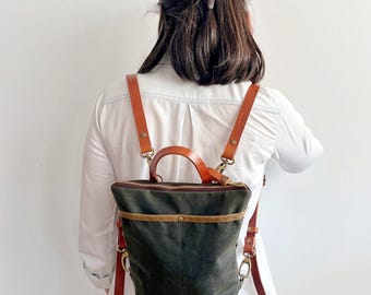 May include: A dark olive green canvas backpack featuring a top zip closure, a horizontal brown leather accent strip, and a top handle. The bag includes adjustable tan leather shoulder straps attached with brass-toned metal swivel clips.