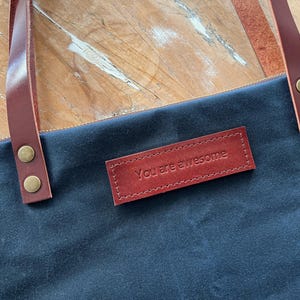 以下が含まれることがあります： 真鍮色の金属製リベットで固定された茶色のレザーハンドルが付いたダークブルーのトートバッグ。前面には長方形の茶色のレザーパッチが縫い付けられており、シンプルで明確なフォントで「You are awesome」というテキストが刻印されています。
