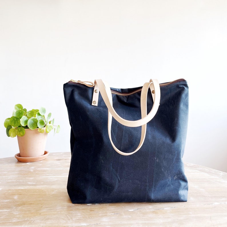 The bag sits on a table. It´s made in blue waxed canvas with natural leather handles.