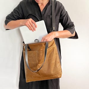 以下が含まれることがあります： ダークブラウンのレザー製ストラップと上部のジッパー開閉を備えた、タンカラーのワックスキャンバス製トートバッグ。バッグには外ポケットがあり、シルバーのノートパソコンを中に入れている様子が示されています。