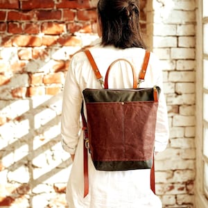 Puede incluir: Una mochila marrón y negra con correas de cuero y un asa superior. La mochila tiene un bolsillo frontal y un cierre de cremallera. La persona lleva una camisa blanca y vaqueros azules.