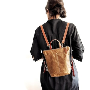 May include: A brown leather backpack with a zipper closure and adjustable straps. The backpack is worn by a person with dark hair.