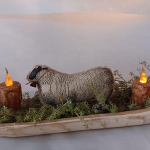 May include: A fabric sheep figurine with a cream and black textured pattern, positioned in a long, shallow, light-colored wooden dough bowl. The bowl is filled with green faux foliage and two brown, textured, battery-operated pillar candles.