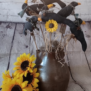 May include: A dark brown ceramic pitcher filled with fabric crow ornaments on sticks, each wearing a small checkered scarf. A single yellow sunflower is attached to one stick, and a cluster of yellow sunflowers sits at the base of the pitcher.