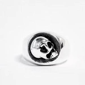 May include: A polished silver-tone ring featuring a raised, three-dimensional skull motif centered on a circular face. The skull design includes detailed eye sockets and nasal cavity, set against a smooth, rounded band with a reflective finish.