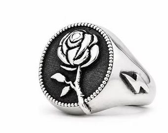 May include: A silver signet ring featuring a circular face with a raised rose design against a dark, textured background. The circular face is bordered by a beaded edge. A small, polished lightning bolt emblem is engraved on the side of the band.