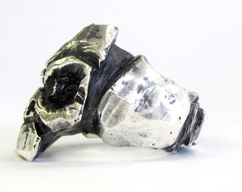 Può includere: Un anello d'argento con un designo testurizzato e astratto. L'anello è fatto di metallo e ha un aspetto ruvido e organico.