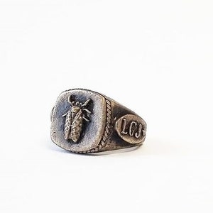 May include: A silver ring with a square face featuring a detailed insect design and the initials "LCJ" engraved on the side.