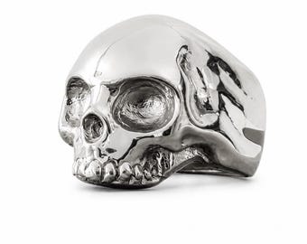 Può includere: Un anello color argento lucido a forma di teschio umano. L'anello presenta una superficie liscia e altamente riflettente con rientranze dettagliate per le orbite, la cavità nasale e i denti, creando un design anatomico realistico.
