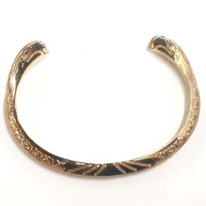 May include: A gold-toned metal cuff bracelet featuring an open design. The surface is decorated with intricate embossed scrollwork patterns along the sides and a series of dark, recessed radial lines at the center of the band's exterior.
