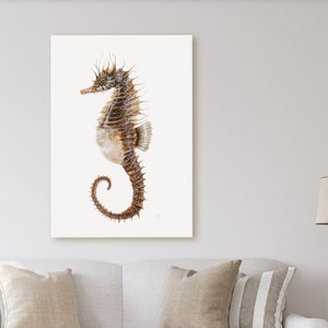 May include: A framed print featuring a detailed watercolor illustration of a seahorse. The seahorse is depicted in shades of brown and tan, with a curled tail and spiky dorsal fin. The background is a soft, off-white color, creating a serene coastal theme.