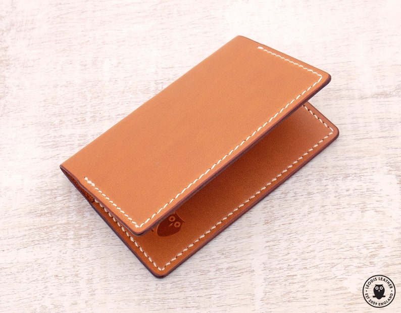 May include: A brown leather card holder with white stitching. The card holder has a small owl logo embossed on the front.
