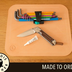 May include: A light brown leather work mat with rounded corners. The mat has a small, embossed owl logo in the lower left corner. The mat features a set of colorful hex keys, a small tube of glue, and a folding knife with a brown handle.