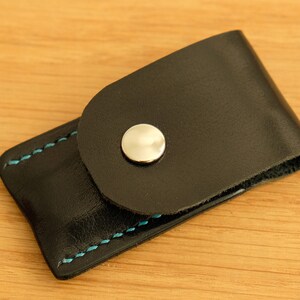 May include: A black leather case with a snap closure and blue stitching. The case has a silver button on the front.
