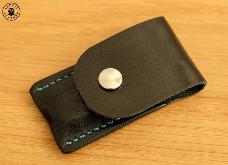 May include: A black leather case with a snap closure and blue stitching. The case has a silver button on the front.