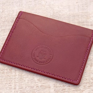 May include: A burgundy leather card holder with a pink stitch detail. The card holder has a single pocket and a debossed logo that reads "LODDIS LEATHER EST 2002 ENGLAND".