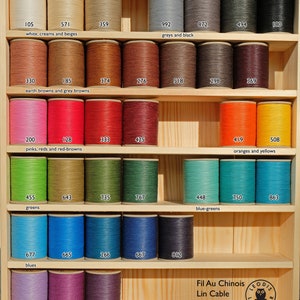 May include: A wooden shelf display of spools of waxed linen thread in various colours, including white, cream, beige, brown, red, orange, yellow, green, blue, purple, and black. The spools are labelled with numbers and colour categories. The text "Fil Au Chinois Lin Cable" and a logo are visible at the bottom of the image.