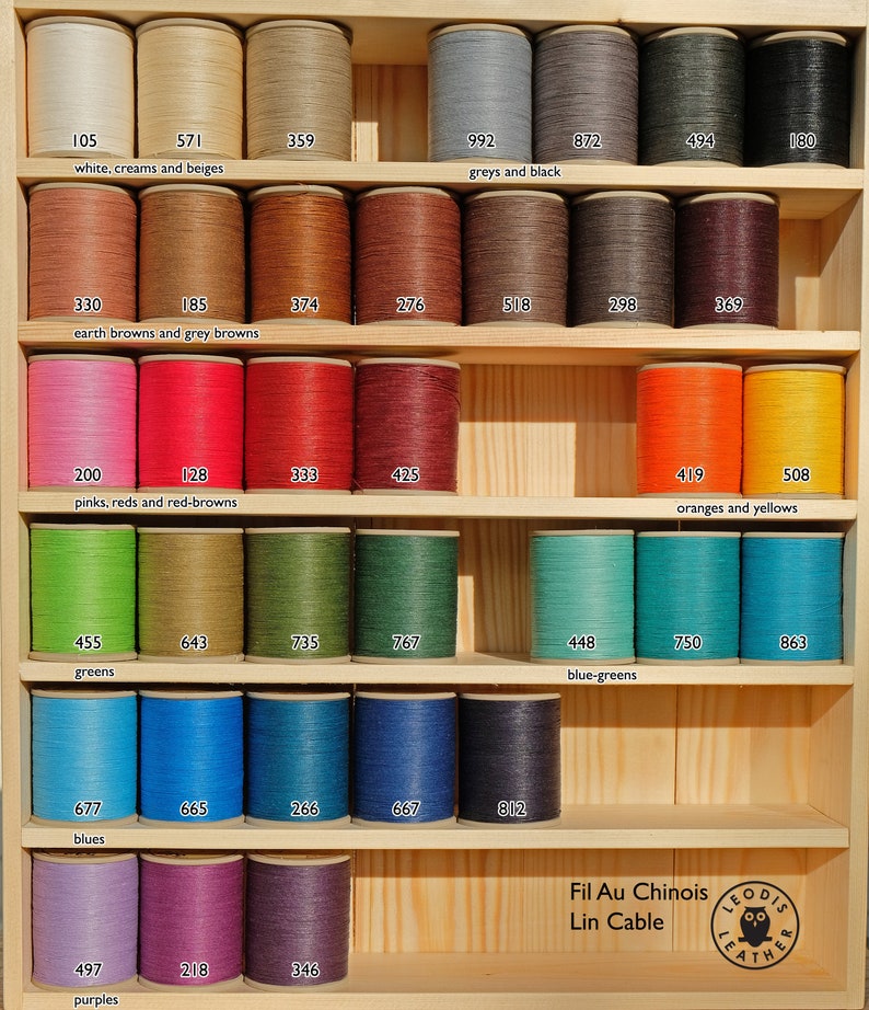 May include: A wooden shelf display of spools of waxed linen thread in various colours, including white, cream, beige, brown, red, orange, yellow, green, blue, purple, and black. The spools are labelled with numbers and colour categories. The text "Fil Au Chinois Lin Cable" and a logo are visible at the bottom of the image.