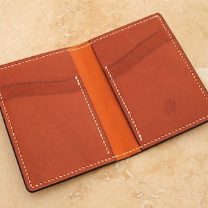 May include: A brown leather wallet with white stitching and an orange interior. The wallet has multiple card slots and a small pocket for cash. The wallet is open and the inside is visible.