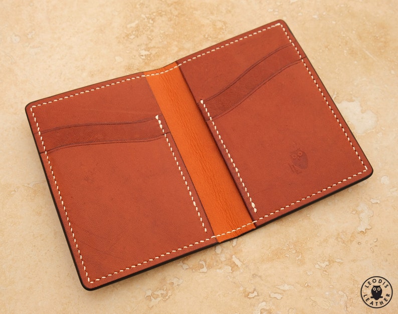 May include: A brown leather wallet with white stitching and an orange interior. The wallet has multiple card slots and a small pocket for cash. The wallet is open and the inside is visible.