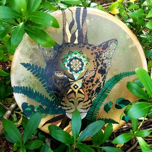 Könnte beinhalten: Eine handbemalte Trommel mit einer detaillierten Illustration eines Jaguar-Gesichts. Der Jaguar hat ein geometrisches Muster auf seiner Stirn und ist von grünem Laub umgeben.