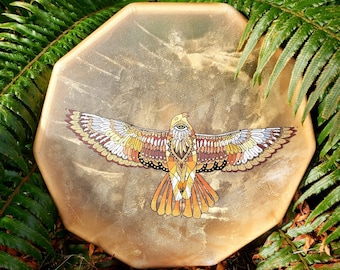 May include: A round, tan-colored drum with a detailed, colorful illustration of a hawk with outstretched wings. The hawk has a large, single eye in the center of its head.