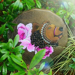 May include: A brown leather drum with a painted design of a bear wearing a beaded headdress and surrounded by pink flowers.