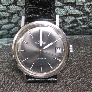 May include: A silver Glycine automatic wristwatch with a black leather band and a gray face. The watch has a date window at the 3 o'clock position and the words "Glycine Automatic" printed on the face.