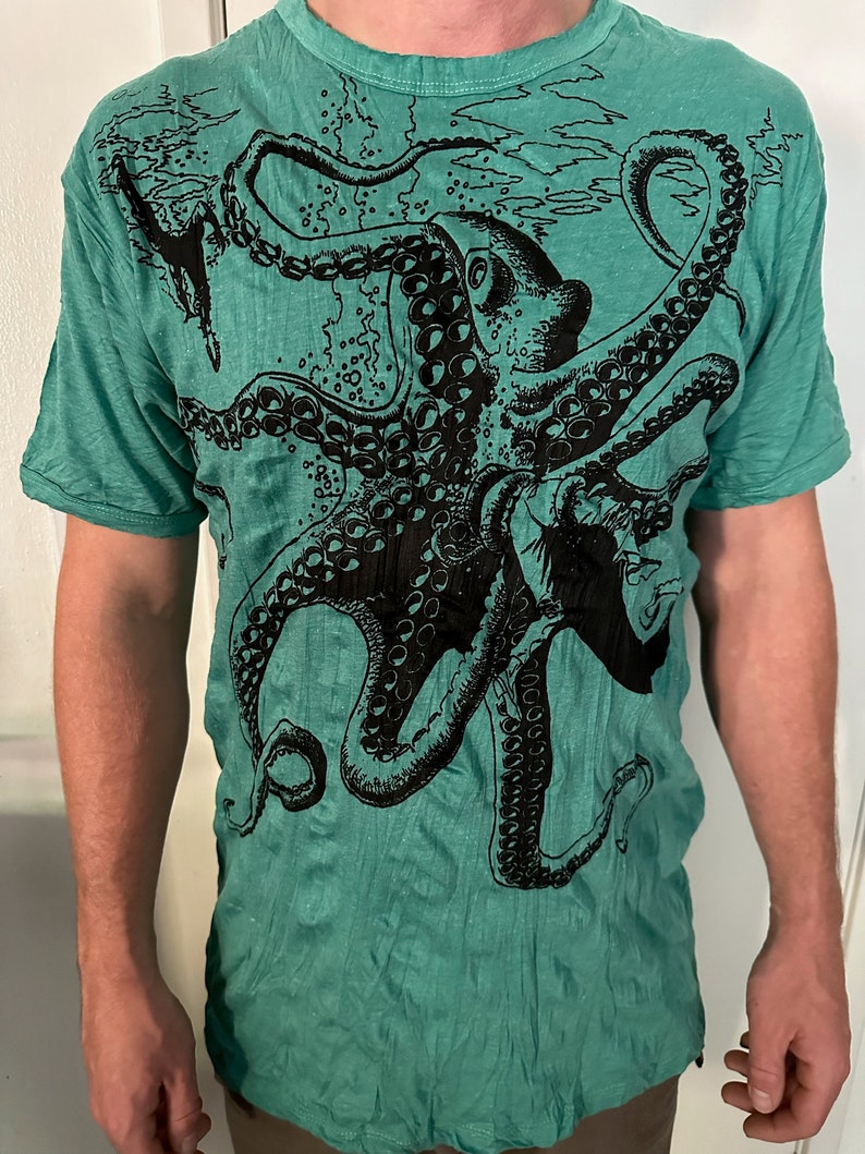 May include: A short-sleeved t-shirt in a vibrant teal green color featuring a large, detailed black ink-style graphic print of an octopus. The design covers the front of the shirt, showcasing the creature's tentacles and suction cups.