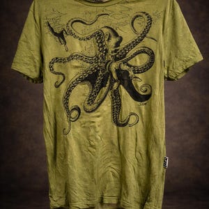 May include: An olive green short-sleeve t-shirt featuring a large black ink-style illustration of an octopus with tentacles spread across the chest. The design includes a small diver figure and subtle underwater textures. A small white tag is on the side.
