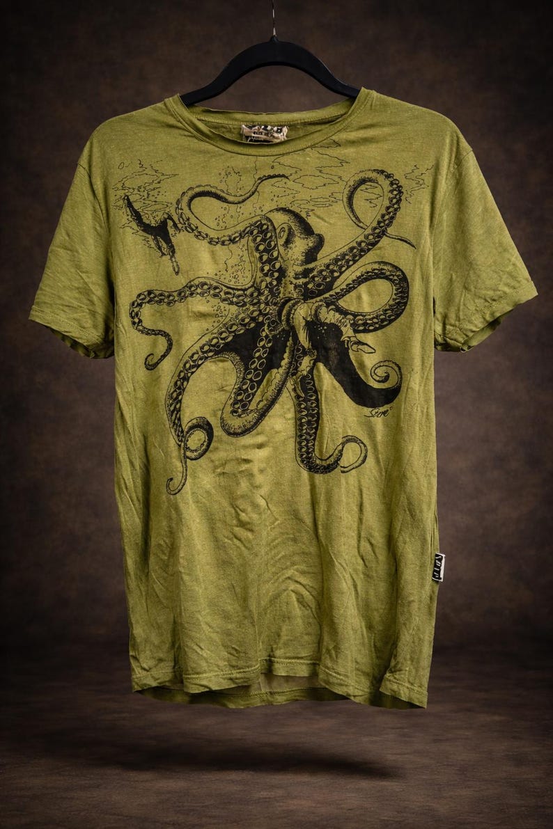 May include: An olive green short-sleeve t-shirt featuring a large black ink-style illustration of an octopus with tentacles spread across the chest. The design includes a small diver figure and subtle underwater textures. A small white tag is on the side.
