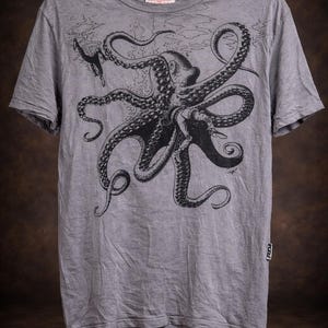May include: A heather gray short-sleeve t-shirt featuring a large black ink-style illustration of an octopus with long tentacles and a small diver figure. The shirt has a crew neckline and a small rectangular brand tag on the lower left side seam.