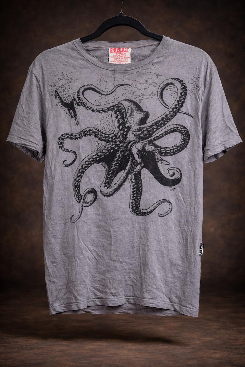 May include: A heather gray short-sleeve t-shirt featuring a large black ink-style illustration of an octopus with long tentacles and a small diver figure. The shirt has a crew neckline and a small rectangular brand tag on the lower left side seam.
