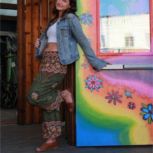 May include: A woman wearing a white crop top, a denim jacket, and green printed pants with a brown waistband. She is standing next to a colorful door with flowers painted on it.
