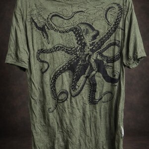 May include: An olive green short-sleeve t-shirt featuring a large black ink-style illustration of an octopus with long, detailed tentacles extending across the chest. The fabric has a heathered texture, and the design includes subtle water-like lines.