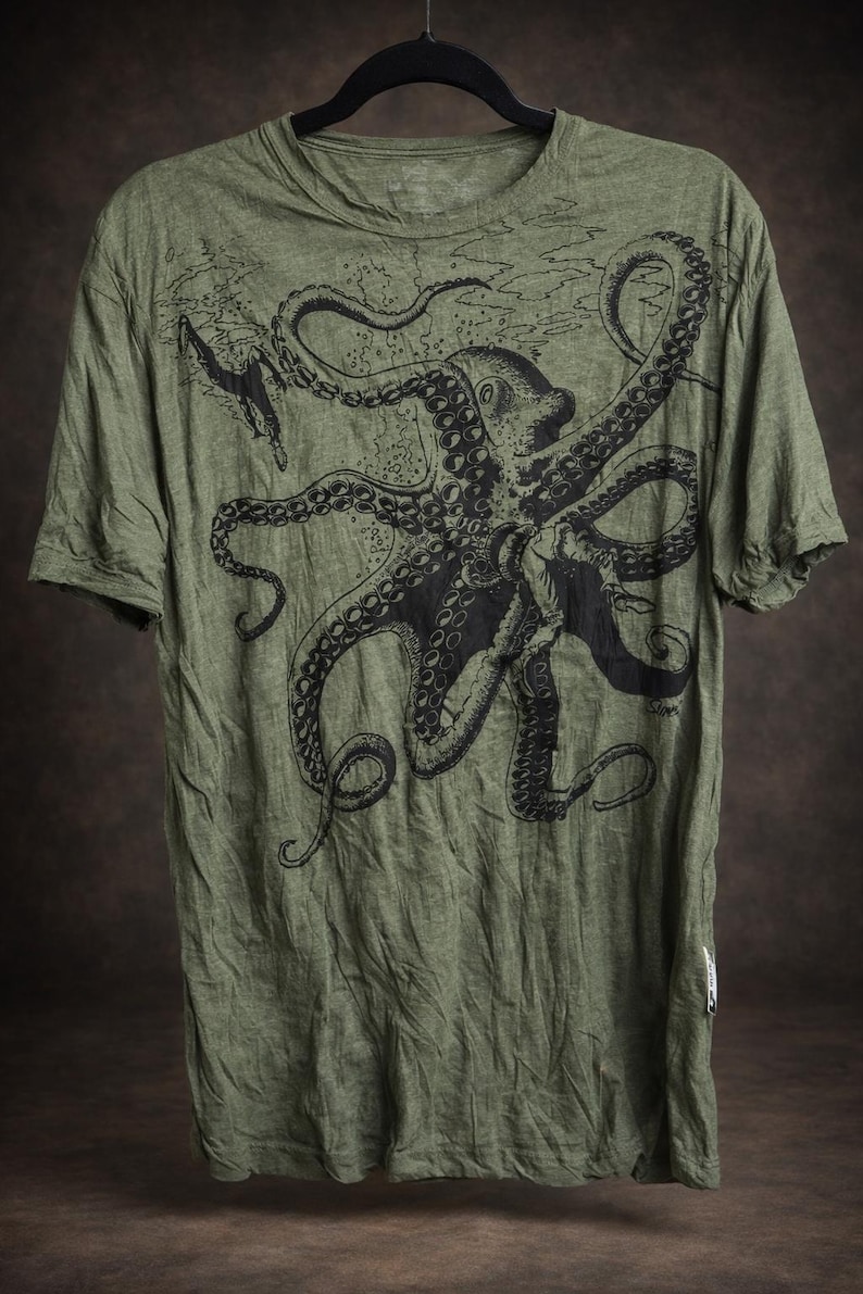 May include: An olive green short-sleeve t-shirt featuring a large black ink-style illustration of an octopus with long, detailed tentacles extending across the chest. The fabric has a heathered texture, and the design includes subtle water-like lines.