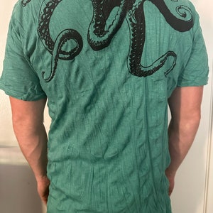 May include: A short-sleeved t-shirt in a heathered green fabric featuring a large, black graphic print of an octopus across the upper back. The design displays the octopus head and multiple tentacles extending across the shoulder area.