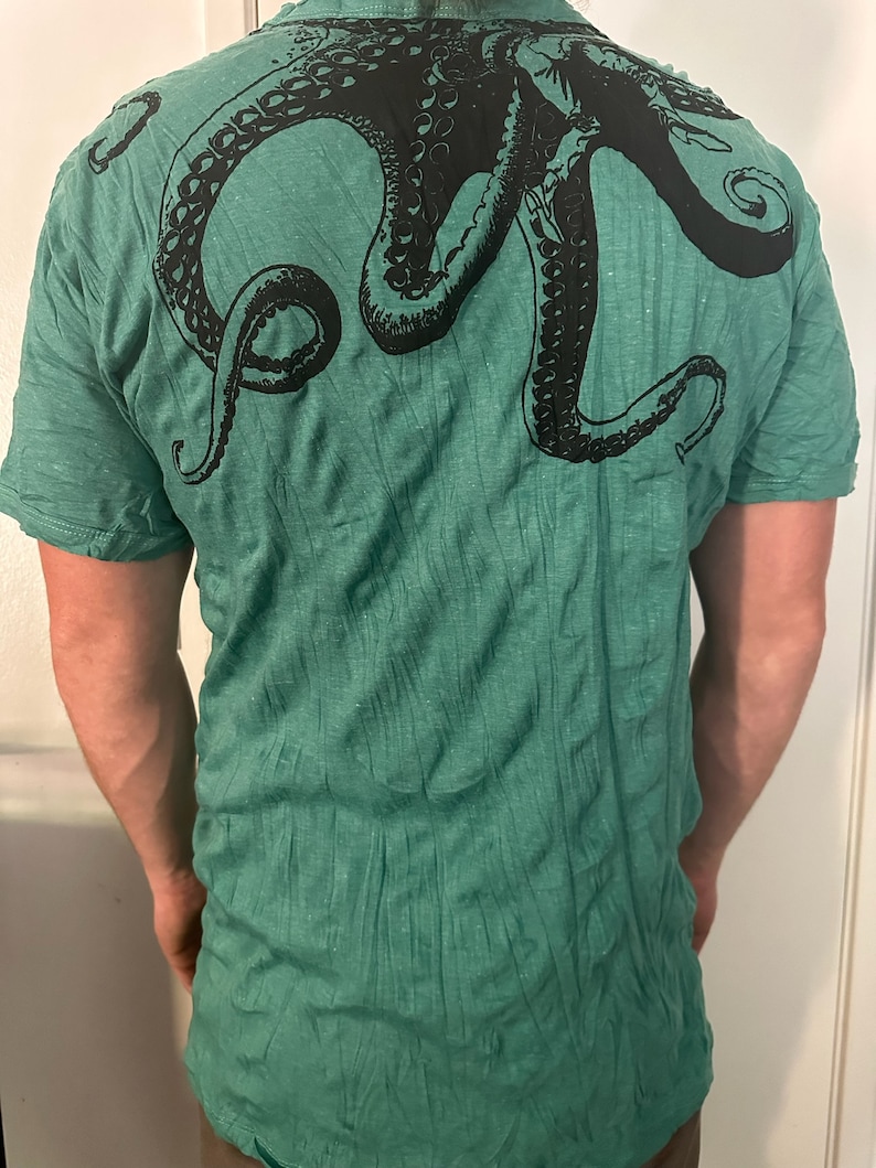 May include: A short-sleeved t-shirt in a heathered green fabric featuring a large, black graphic print of an octopus across the upper back. The design displays the octopus head and multiple tentacles extending across the shoulder area.