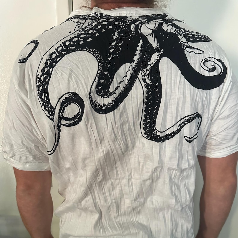 May include: A white short-sleeved t-shirt featuring a large, detailed black ink-style graphic of an octopus printed across the upper back. The design showcases the creature's head and multiple long, curling tentacles with visible suction cups.