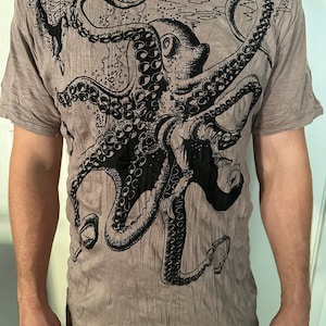 May include: A short-sleeved, crew-neck t-shirt in a muted taupe color featuring a large, detailed black ink-style illustration of an octopus. The graphic covers the front of the garment, showcasing the creature's tentacles and suckers.