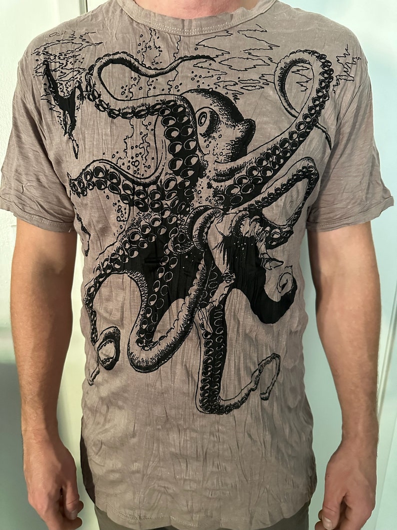May include: A short-sleeved, crew-neck t-shirt in a muted taupe color featuring a large, detailed black ink-style illustration of an octopus. The graphic covers the front of the garment, showcasing the creature's tentacles and suckers.