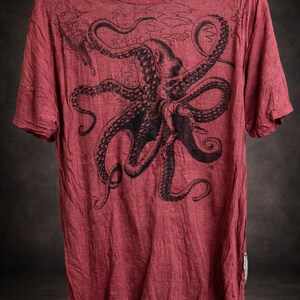 May include: A short-sleeved, crew-neck t-shirt in a faded red color. The front features a large, detailed black ink-style illustration of an octopus with its tentacles spread across the chest area. The fabric has a slightly textured, worn appearance.